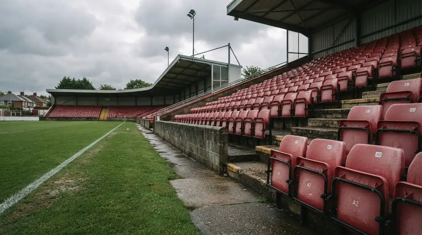 Rzędy pustych plastikowych krzesełek małego stadionu widziane z poziomu murawy pod pochmurnym niebem.