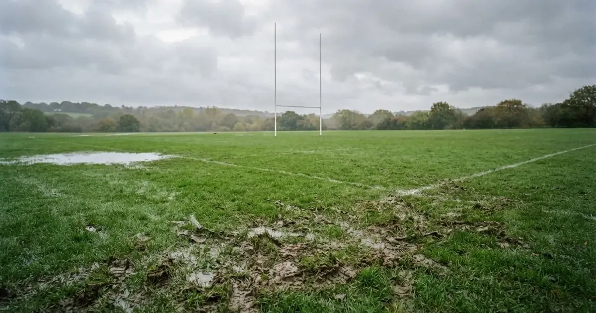 Boisko do rugby ze słupami bramki w oddali pod pochmurnym niebem, mokra trawa i ślady butów.