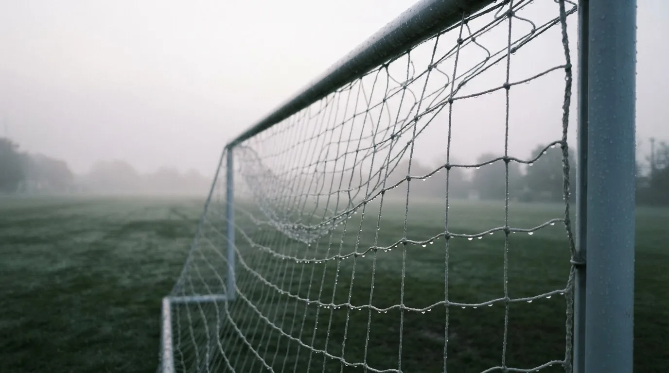 Siatka bramki piłkarskiej w kroplach deszczu, ujęcie z boku o szarym poranku.
