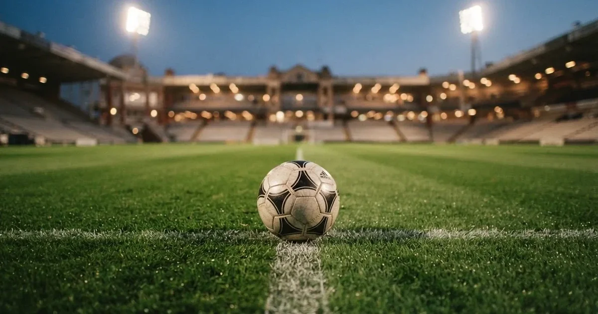 Piłka nożna leży na środku świeżo skoszonej murawy, w tle pusty stadion w świetle reflektorów wczesnym wieczorem.